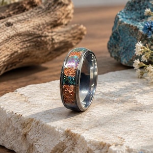 May include: A metallic ring featuring a central inlay of iridescent, multi-colored crushed flakes in shades of orange, green, and blue. The band has a polished, reflective silver-toned finish and a smooth, rounded profile with a wide, flat band design.