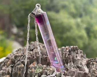 Op de afbeelding: Een zilveren ketting met een hanger van paarse en iriserende hars. De hanger is lang en dun cilindrisch gevormd en heeft bovenaan een zilveren lus.