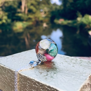 Puede incluir: Un collar de plata con un colgante redondo lleno de resina colorida y pequeñas conchas. El colgante tiene un diseño decorativo de plata alrededor de la parte superior.