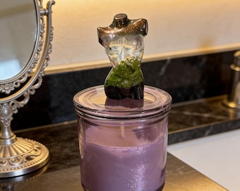 May include: A lavender-colored candle in a clear glass jar, topped with a decorative torso-shaped object. The candle has a glittery appearance. The torso is clear with a brown glitter top and green moss at the bottom.
