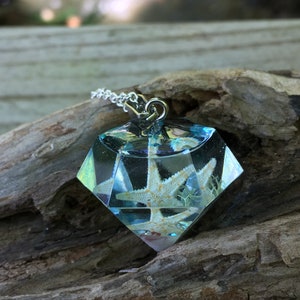 Puede incluir: Un collar con un colgante en forma de diamante que contiene una estrella de mar blanca y otros elementos marinos en una resina transparente. El colgante está en una cadena de plata.