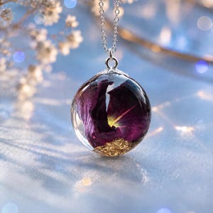 May include: A spherical resin pendant necklace featuring a deep purple dried flower petal and gold leaf flakes suspended inside the clear orb. The pendant is attached to a thin silver-toned metal chain with a decorative silver cap at the top.