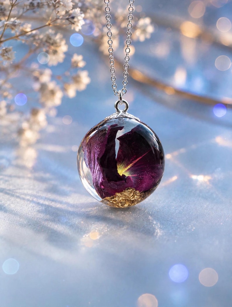 May include: A spherical resin pendant necklace featuring a deep purple dried flower petal and gold leaf flakes suspended inside the clear orb. The pendant is attached to a thin silver-toned metal chain with a decorative silver cap at the top.