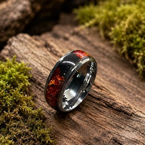May include: A metallic ring featuring a polished silver-toned band with a wide inlay of black material embedded with shimmering, irregular red and orange flakes that catch the light, creating a distinct contrast against the smooth metal edges.