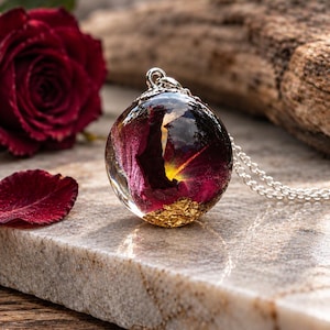 May include: A spherical resin pendant necklace featuring a preserved dark red rose petal and gold leaf flakes suspended inside the clear orb. The pendant is attached to a silver-toned metal chain with a small decorative bail at the top.