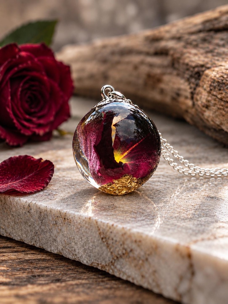 May include: A spherical resin pendant necklace featuring a preserved dark red rose petal and gold leaf flakes suspended inside the clear orb. The pendant is attached to a silver-toned metal chain with a small decorative bail at the top.