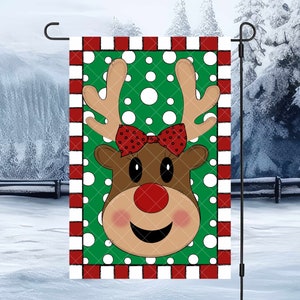 May include: A green and white garden flag with a red and white striped border. The flag features a cartoon reindeer face with a red nose, brown antlers, and a red bow.