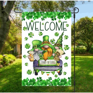 May include: A welcome garden flag featuring three gnomes wearing green hats and holding beer mugs. The gnomes are driving a blue car with the words "Loads of Luck" on the side. The flag is surrounded by green shamrocks.