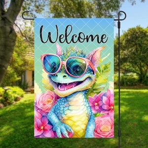 May include: A garden flag with a cartoon dragon wearing pink sunglasses and surrounded by pink, purple, and blue flowers. The flag says "Welcome" in black text.