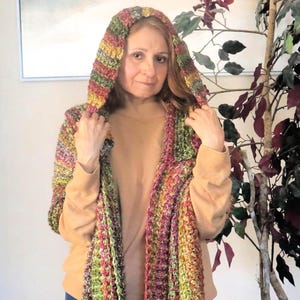 May include: A hooded crochet scarf in a variety of colors, including green, pink, yellow, and orange. The scarf is worn over a tan long-sleeve shirt and blue jeans. The scarf is handmade and appears to be warm and cozy.