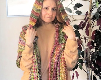 May include: A hooded crochet scarf in a variety of colors, including green, pink, yellow, and orange. The scarf is worn over a tan long-sleeve shirt and blue jeans. The scarf is handmade and appears to be warm and cozy.