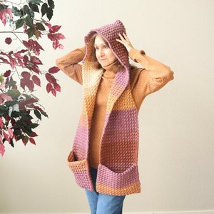 May include: A multi-coloured hooded scarf with pockets. The scarf is made from a soft, chunky yarn and features a gradient of pink, orange, and yellow. The hood is lined with a cream-coloured fabric.