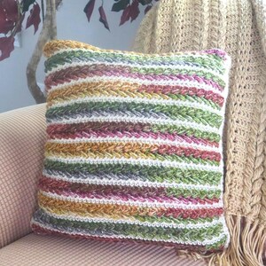 May include: A colorful, striped decorative pillow with a textured, woven design. The pillow features horizontal stripes in shades of green, purple, white, yellow, and brown. A beige crocheted blanket with fringe is draped behind the pillow.