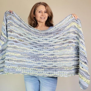May include: A light blue, gray, and yellow crocheted shawl with a wavy pattern. The open weave design creates a delicate, airy look. The shawl is held up, showcasing its size and intricate details. A cozy, handmade accessory.