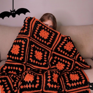 Könnte beinhalten: Eine Frau versteckt sich unter einer schwarzen und orangefarbenen Häkeldecke auf einem beigen Sofa. Die Decke hat ein sich wiederholendes Muster aus schwarzen und orangen Quadraten. An der Wand hinter dem Sofa hängt eine schwarze Fledermaus und auf dem Boden befindet sich eine schwarze Katzensilhouette.