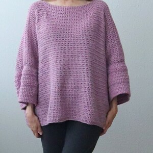 May include: A loose-fitting, crocheted sweater in a pale purple color. The sweater has a wide, boat neck and long sleeves with a slightly flared cuff.