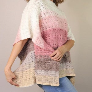 May include: A crocheted poncho with a gradient of colors from white to pink to beige. The poncho has a loose fit and a wide neckline. The model is wearing the poncho with blue jeans.