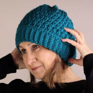 Può includere: Una donna indossa un berretto di lana color turchese lavorato a uncinetto. Indossa un maglione nero con un motivo a rombi rosso e grigio.