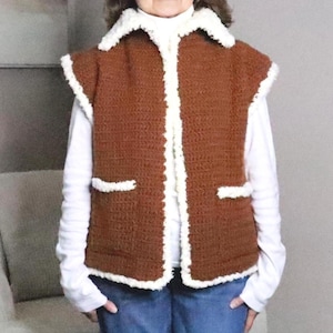 May include: A sleeveless brown crochet vest featuring a white faux-shearling trim along the collar, front opening, and two front pockets. The vest is worn over a long-sleeved white shirt and blue jeans, showcasing a textured, knit construction.