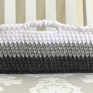 May include: A white, gray, and dark gray crocheted basket with a handle. The basket is made with a textured stitch and has a rectangular shape.