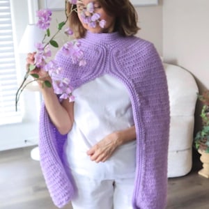 Puede incluir: Una capa de ganchillo lila con mangas largas. La capa tiene un cuello alto y redondeado y un patrón de puntada texturizado. Las mangas son largas y anchas. La capa se lleva sobre una camiseta blanca y pantalones blancos. La capa es de color pastel.