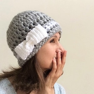 Puede incluir: Un gorro de punto gris con un lazo de punto blanco en un lado.