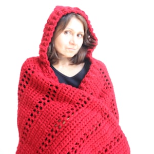 Può includere: Una coperta con cappuccio in crochet rosso con un motivo a pizzo. La coperta è drappeggiata sulle spalle di una persona.