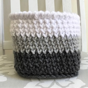 May include: A white, gray, and black knitted storage basket with a textured pattern. The basket is made of yarn and has a round shape.