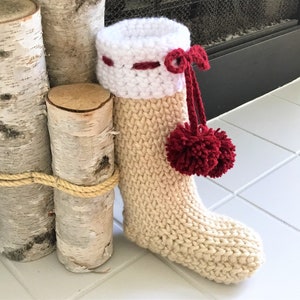 Op de afbeelding: Een beige gebreide kerstsok met een witte gehaakte boord en rode pompoenen. De boord heeft een rood lint en een rode bies.