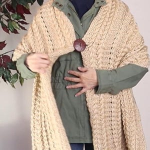 Pode incluir: Um xale de malha bege com um padrão de malha de cabo e um fecho de botão de madeira.