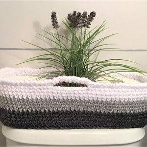 May include: A white, gray, and black crocheted basket with a handle sits on a white toilet tank. The basket is filled with toilet paper rolls and a small potted plant with lavender flowers.