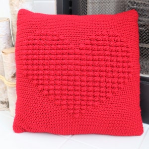 Puede incluir: Almohada roja de ganchillo con un diseño de corazón. La almohada está hecha con un punto texturizado que crea una forma de corazón en relieve.