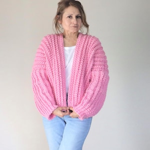 Può includere: Un cardigan rosa acceso a maglia grossa a coste larghe. Il cardigan aperto sul davanti ha maniche lunghe e una vestibilità morbida. La modella indossa una maglietta bianca e jeans blu.