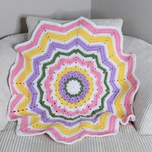 May include: A round crochet blanket featuring a starburst pattern with concentric rings of white, light purple, yellow, green, and pink yarn. The design radiates from a central white circle, creating a textured, wavy edge throughout the piece.