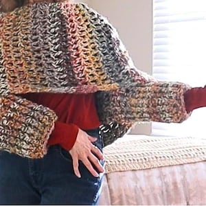Könnte beinhalten: Ein handgefertigter, grob gestrickter Häkelschal in einer Mischung aus Braun-, Orange- und Cremefarben. Der Schal hat weite Ärmel und ein strukturiertes Stichmuster. Das Kleidungsstück wird über einem roten Langarmshirt und einer blauen Jeans getragen.