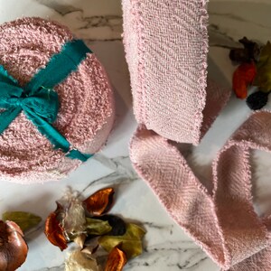 May include: Pink fabric ribbon with a frayed edge. The ribbon is rolled up and there is a smaller roll of pink fabric with a green ribbon tied around it.