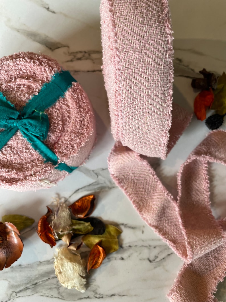 May include: Pink fabric ribbon with a frayed edge. The ribbon is rolled up and there is a smaller roll of pink fabric with a green ribbon tied around it.