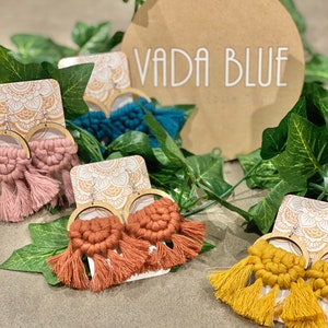 May include: A set of three pairs of handmade macrame earrings with wooden hoops. The earrings are in shades of pink, orange, and yellow with tassels. The earrings are displayed on a white card with the text "VADA BLUE" on a brown circle.