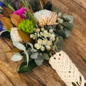 May include: A bouquet of flowers wrapped in a white macrame holder. The bouquet includes eucalyptus, white and pink flowers, and green foliage.