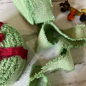 May include: Green fabric ribbon with a frayed edge. The ribbon has a herringbone pattern and is made of a soft, textured material.