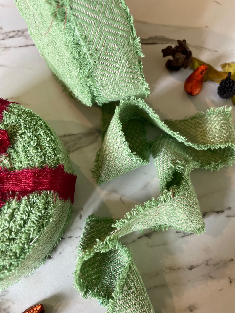 May include: Green fabric ribbon with a frayed edge. The ribbon has a herringbone pattern and is made of a soft, textured material.