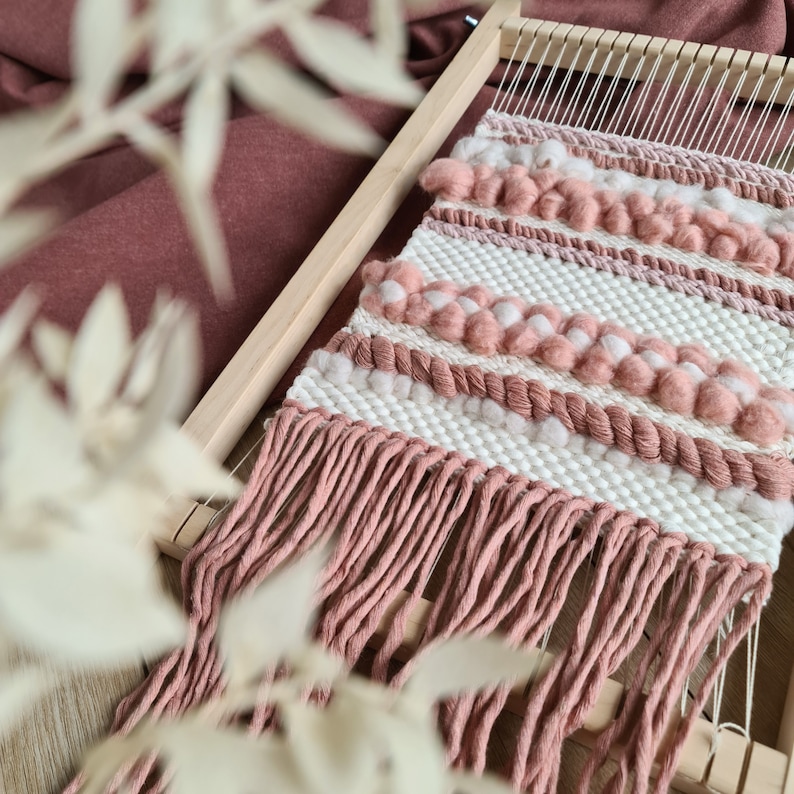 May include: A pink and white woven wall hanging with fringe. The hanging is made with yarn and has a textured pattern.
