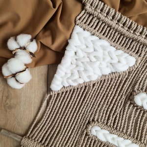 May include: A beige macrame wall hanging with white cotton accents. The wall hanging is made with a thick rope and features a geometric pattern.