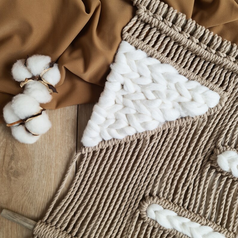 May include: A beige macrame wall hanging with white cotton accents. The wall hanging is made with a thick rope and features a geometric pattern.