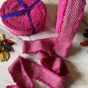 May include: Pink and white woven ribbon with a chevron pattern. The ribbon is frayed on the edges and is rolled up into two coils.
