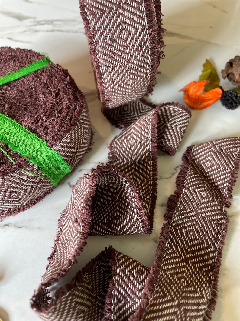 May include: Brown and white woven ribbon with a diamond pattern and frayed edges. The ribbon is rolled up and laid out flat.