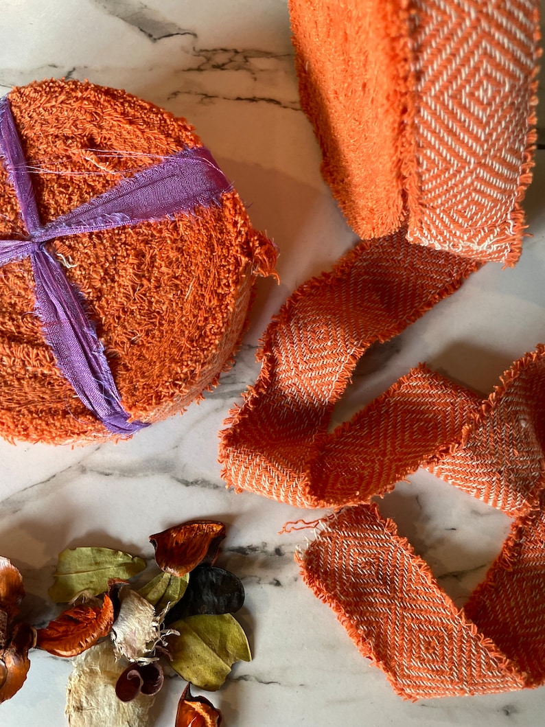 May include: Two orange fabric rolls with a white and orange patterned trim. The rolls are on a white marble surface with dried flower petals.