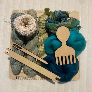 May include: A wooden loom with a variety of yarn and fiber for weaving. The yarn is in shades of green, blue, and white. There is also a wooden comb and a wooden needle.