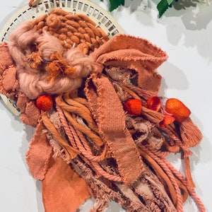 May include: A macrame wall hanging with a woven center and long, fringed strands of yarn in shades of peach and terracotta.