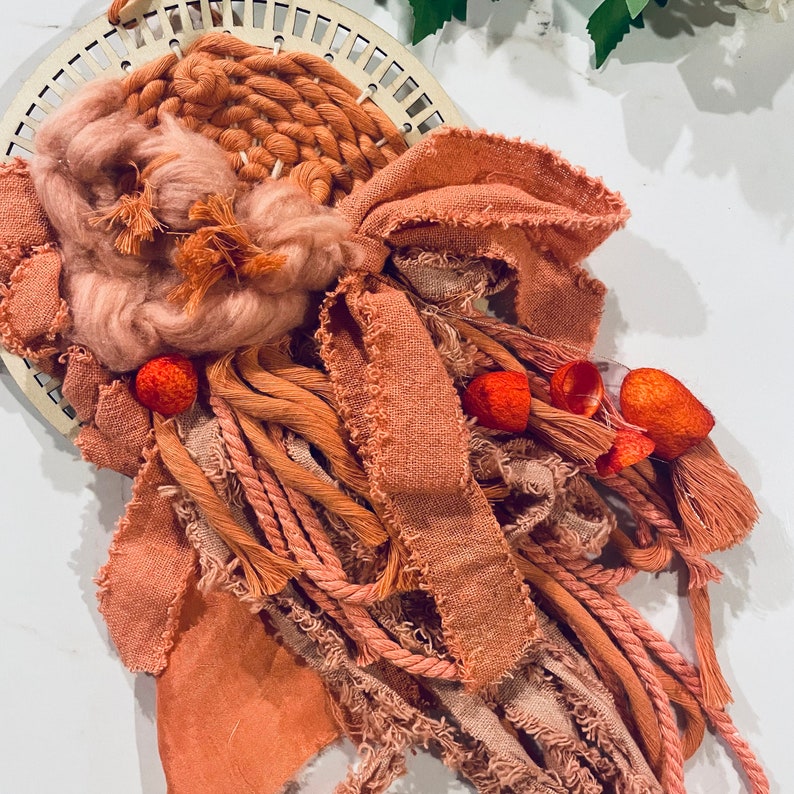 May include: A macrame wall hanging with a woven center and long, fringed strands of yarn in shades of peach and terracotta.